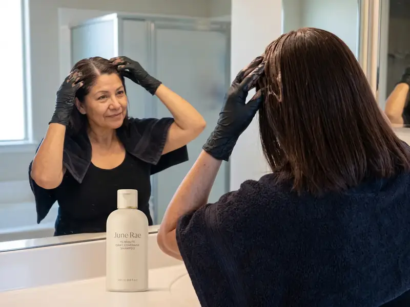 A woman wearing black gloves massages a brown lather into dry hair at her temples and part, the June Rae bottle on the counter
